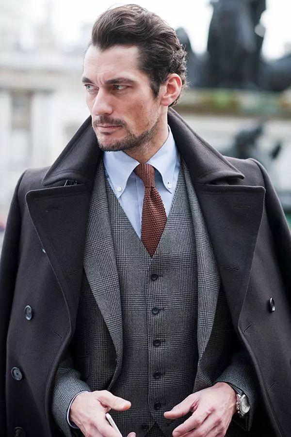 Uomo in completo grigio, cappotto scuro e cravatta, in un ritratto editoriale di ispirazione britannica.